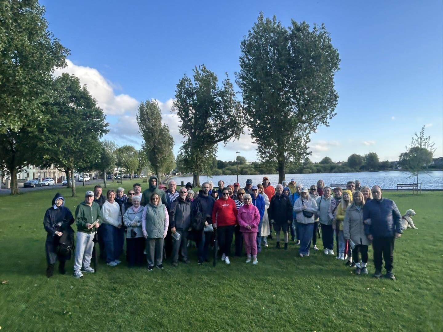 A large group of people standing together on a grassy lakeside on a sunny evening