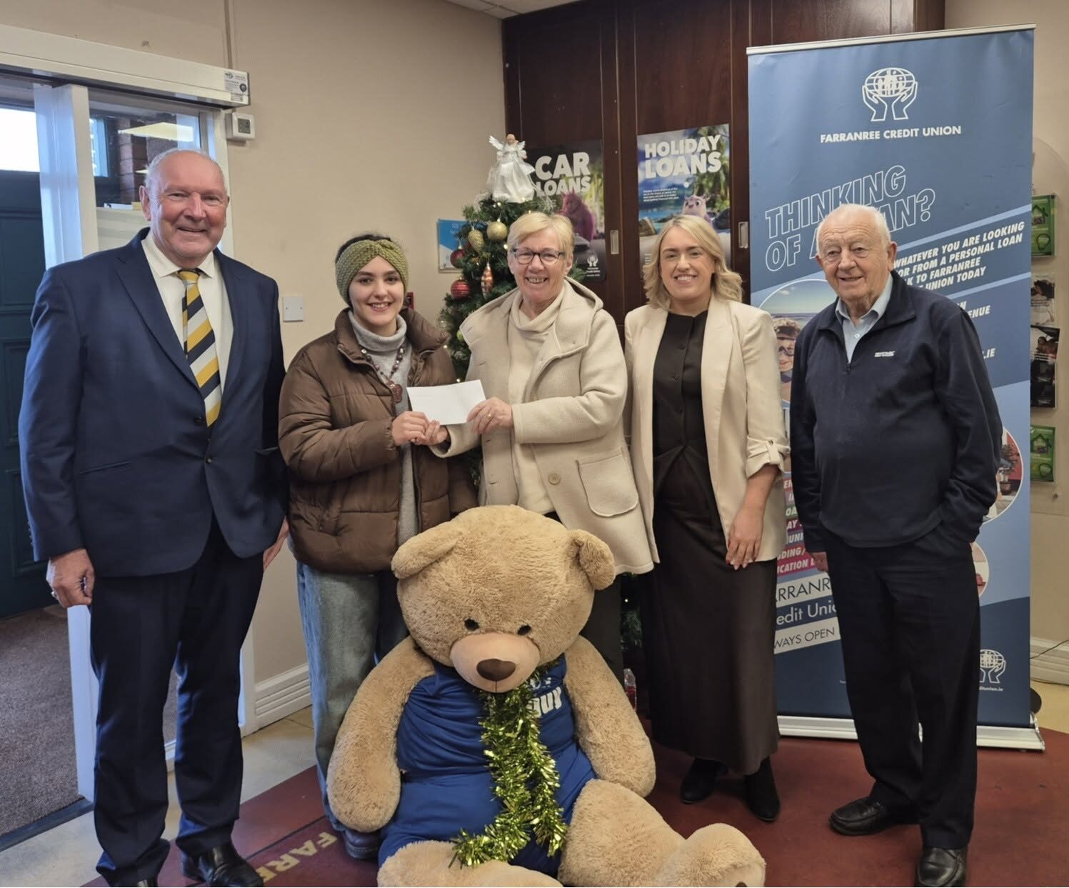 Five people smiling together beside a Christmas tree as an envelope is handed over
